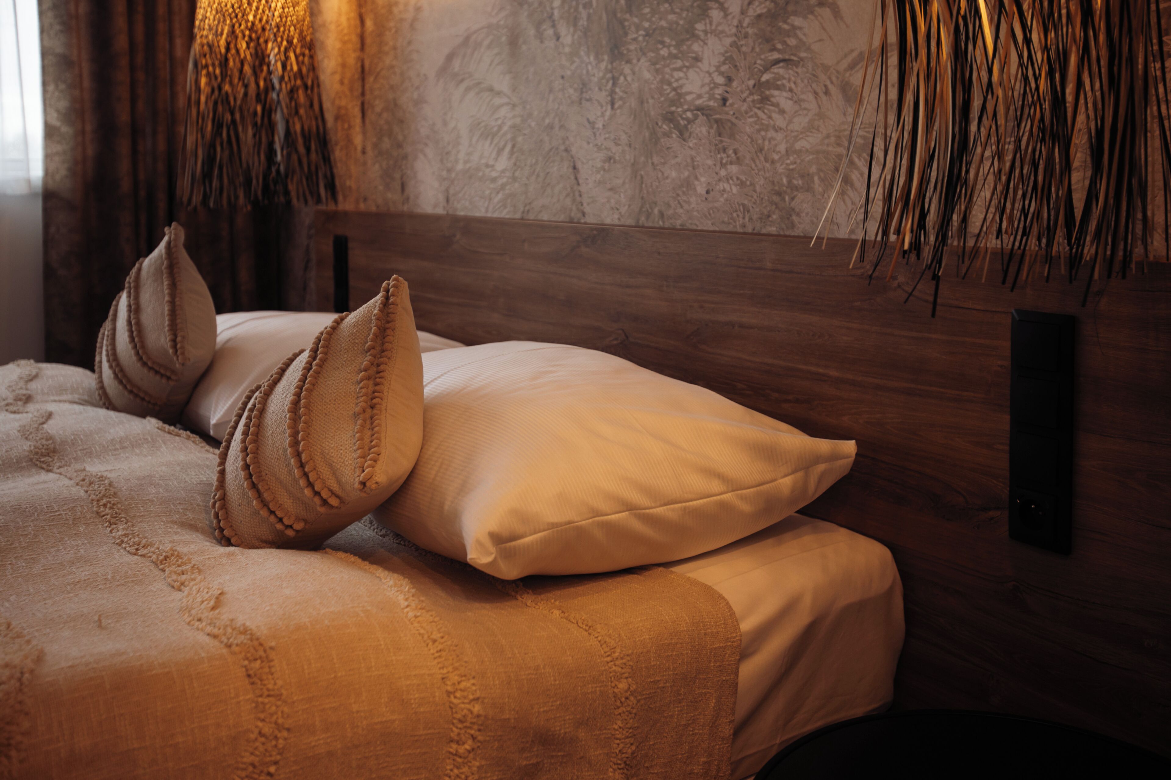 Un lit avec une tête de lit en bois, une literie de couleur beige et un mur texturé agrémenté d’un luminaire suspendu.