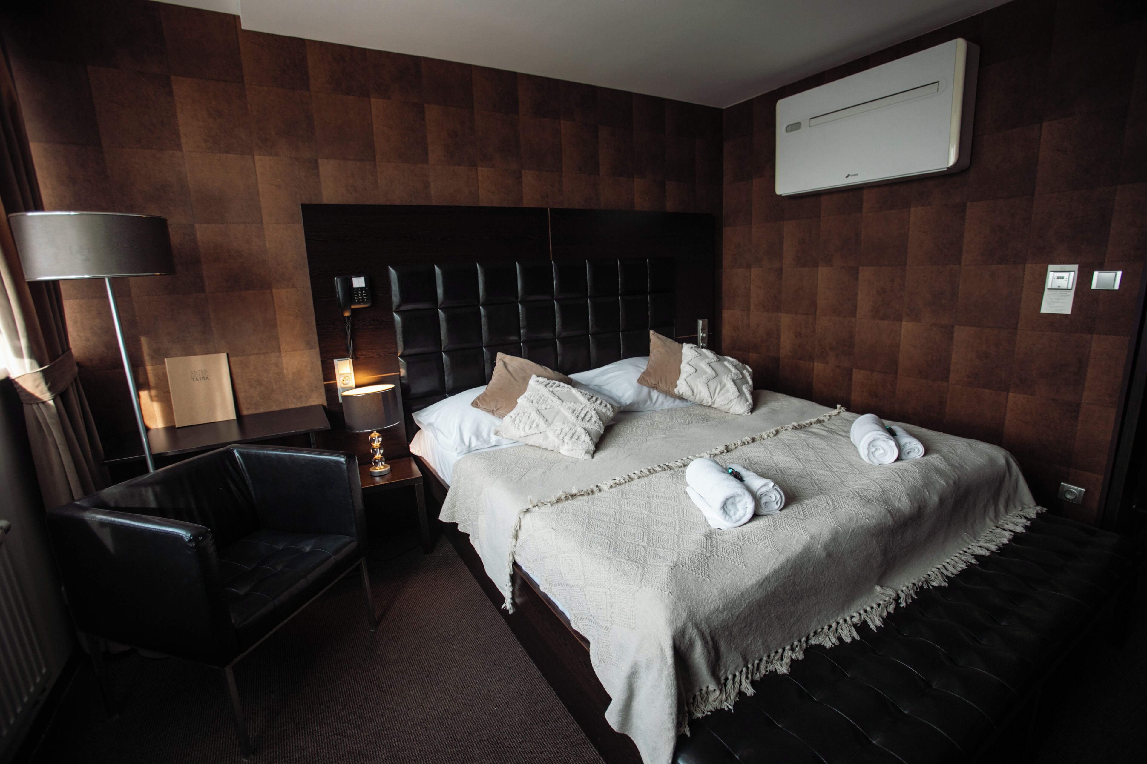 Habitación de hotel con una cama grande, una mesita de noche con lámpara, una silla y un aire acondicionado instalado en la pared.