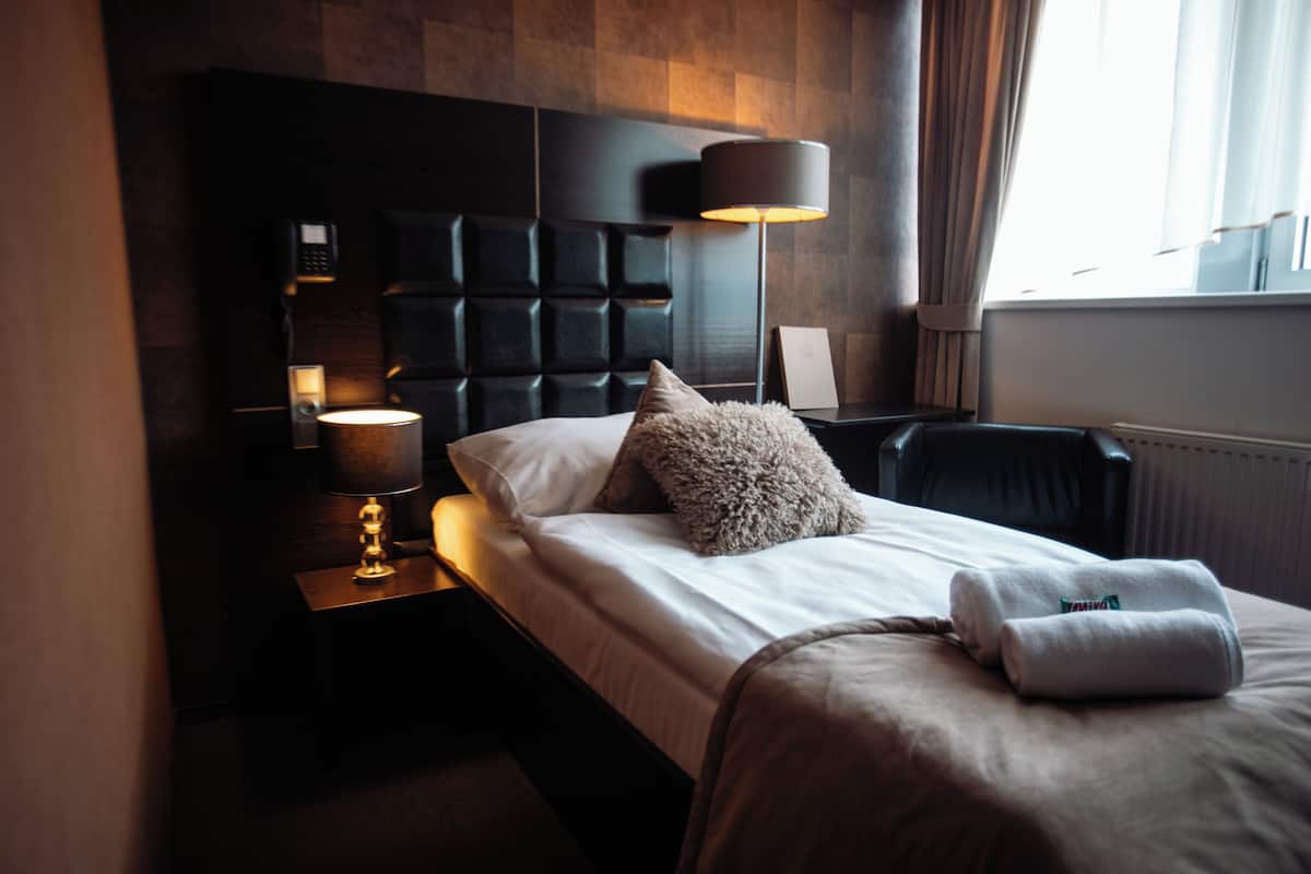 A hotel room with a modern design, featuring a large bed with a headboard, a bedside table with a lamp, and a view of the outside through a window.