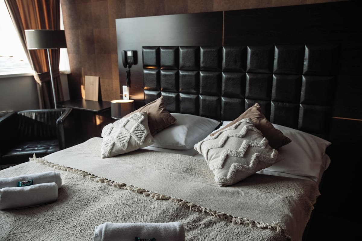 A hotel room with a large bed, a headboard with a textured design, a bedside table with a lamp, and a chair.