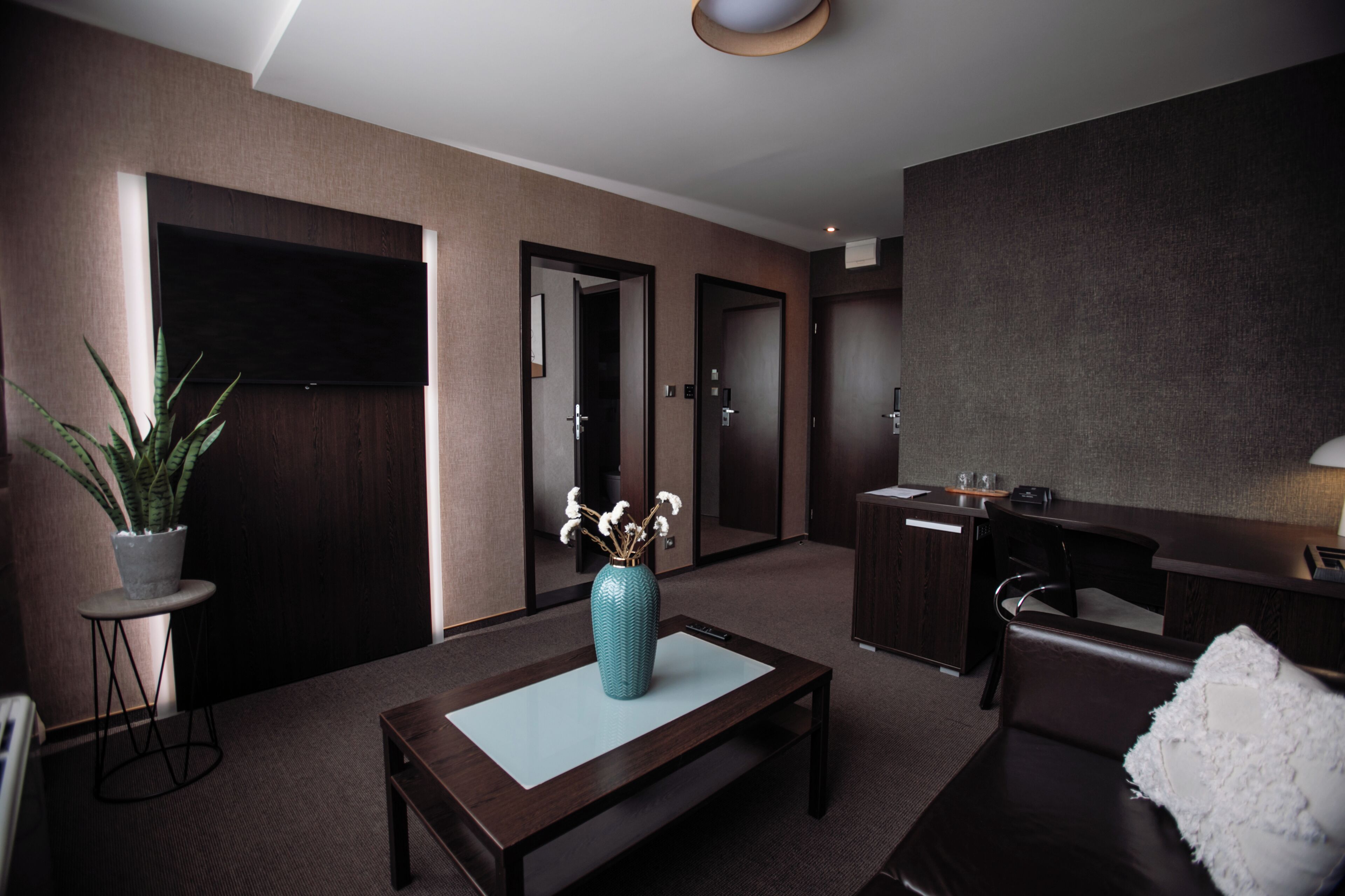 A modern hotel lobby with a flat-screen TV, a reception desk, a seating area with a coffee table, and a potted plant.