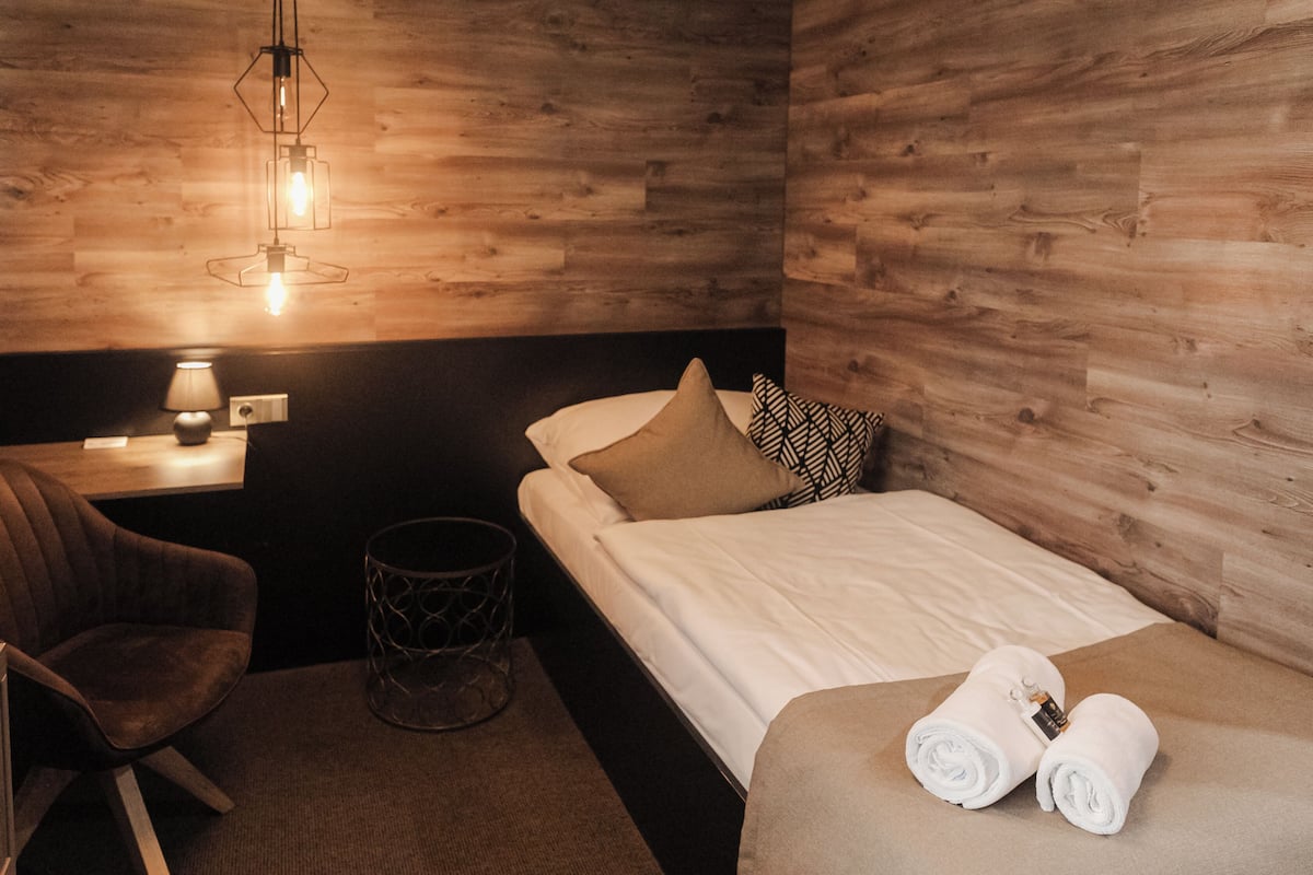 A hotel room with a wooden wall, a bed with white bedding, a chair, a lamp, and a trash bin.