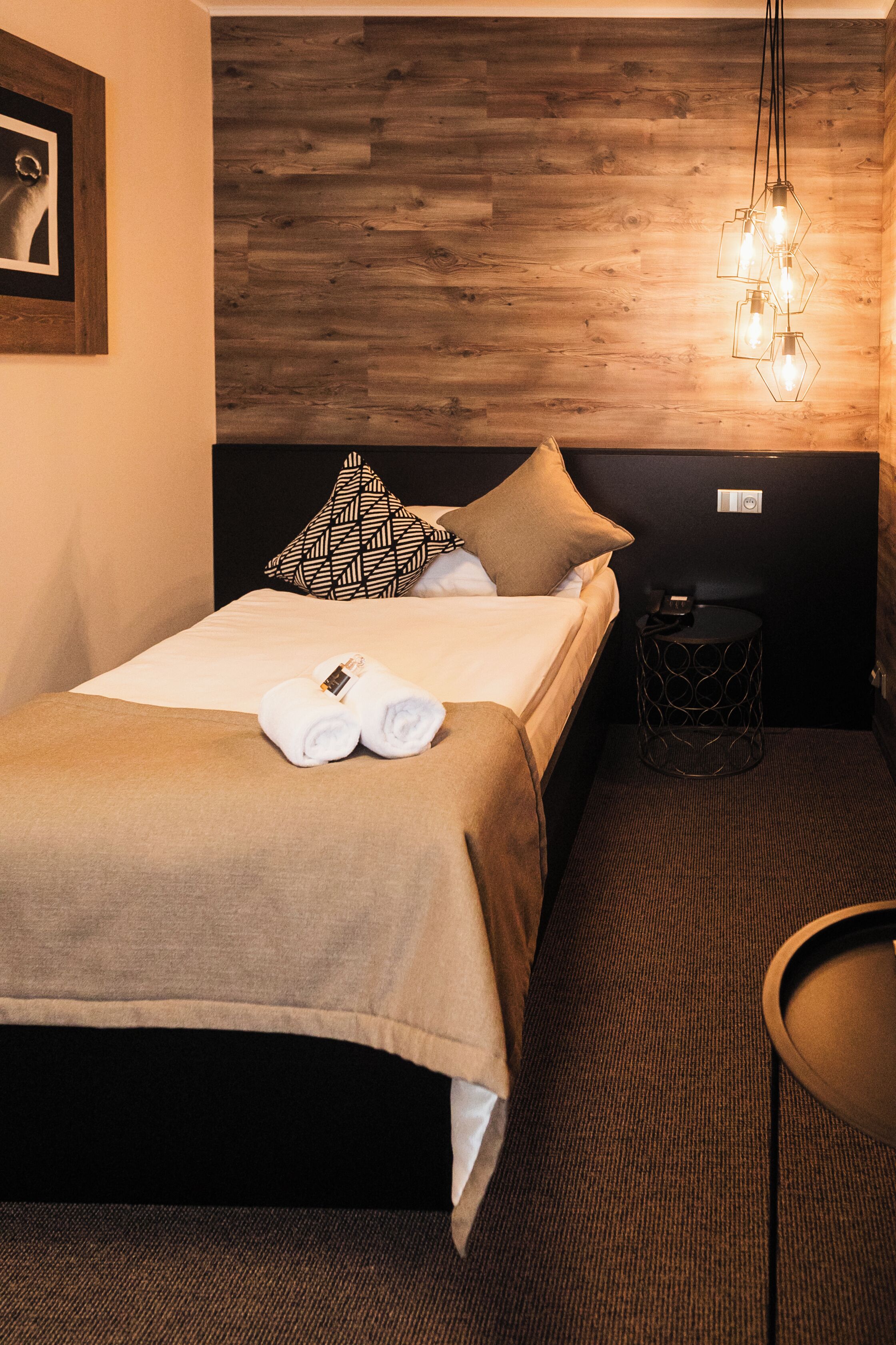 Una cama bien tendida con dos toallas, un cabecero de madera, una lámpara de pared y una mesita redonda con un teléfono y cargador.