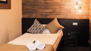 Una cama bien tendida con dos toallas, un cabecero de madera, una lámpara de pared y una mesita redonda con un teléfono y cargador.