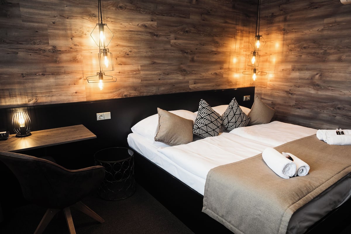 A hotel room with a wooden wall, a bed with a beige blanket, two pillows, a bedside table with a lamp, and a chair.