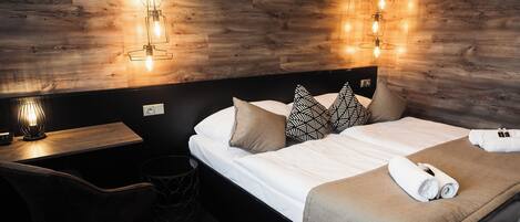 A hotel room with a wooden wall, a bed with a beige blanket, two pillows, a bedside table with a lamp, and a chair.