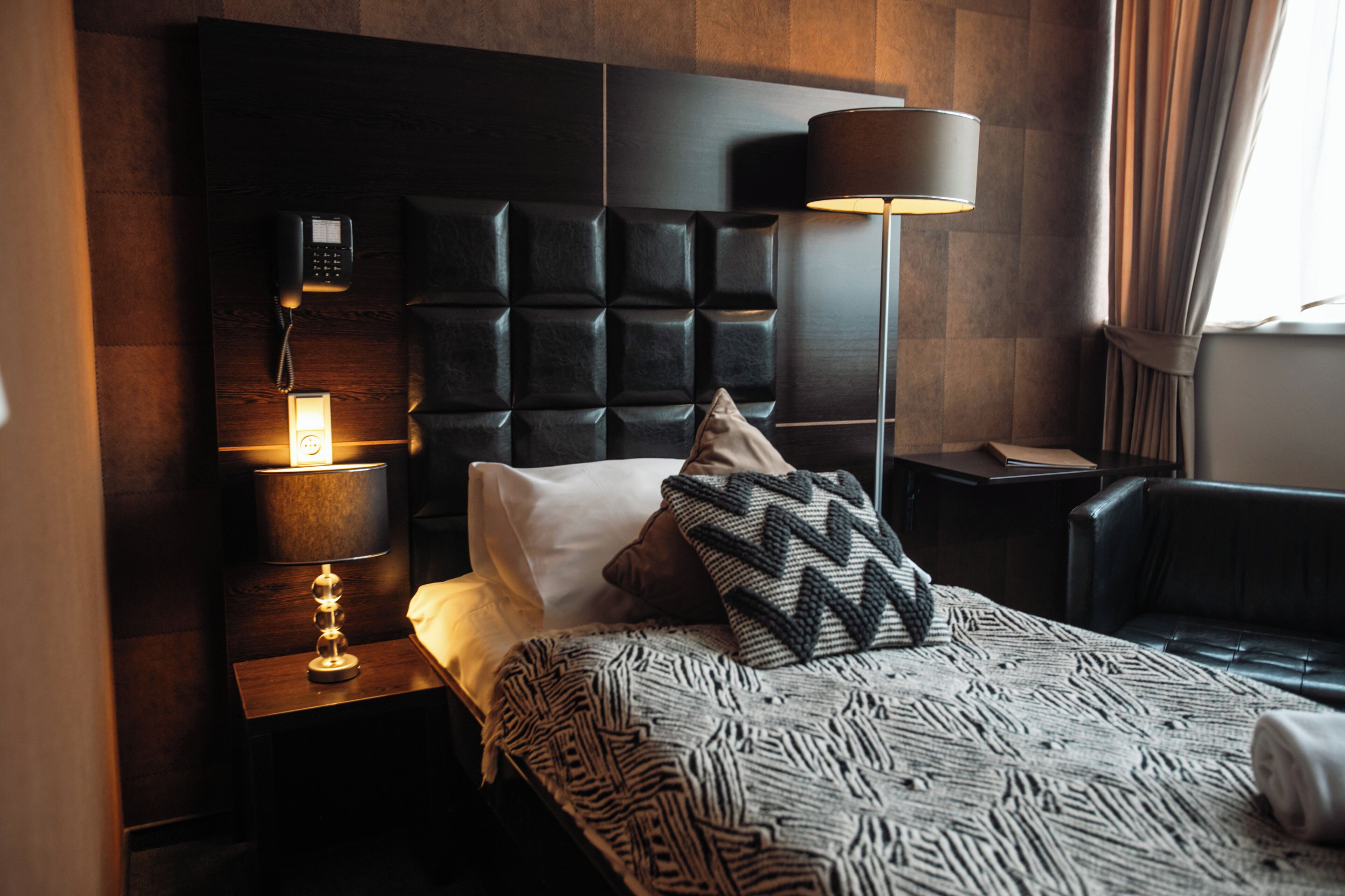 A hotel room with a tufted headboard, a bedside lamp, a telephone, and a patterned throw blanket.