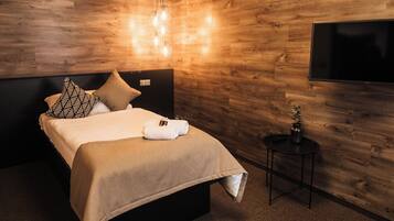 A modern bedroom with a wooden wall, a bed with a beige bedspread, two patterned pillows, a bedside table with a vase, and a mounted TV.
