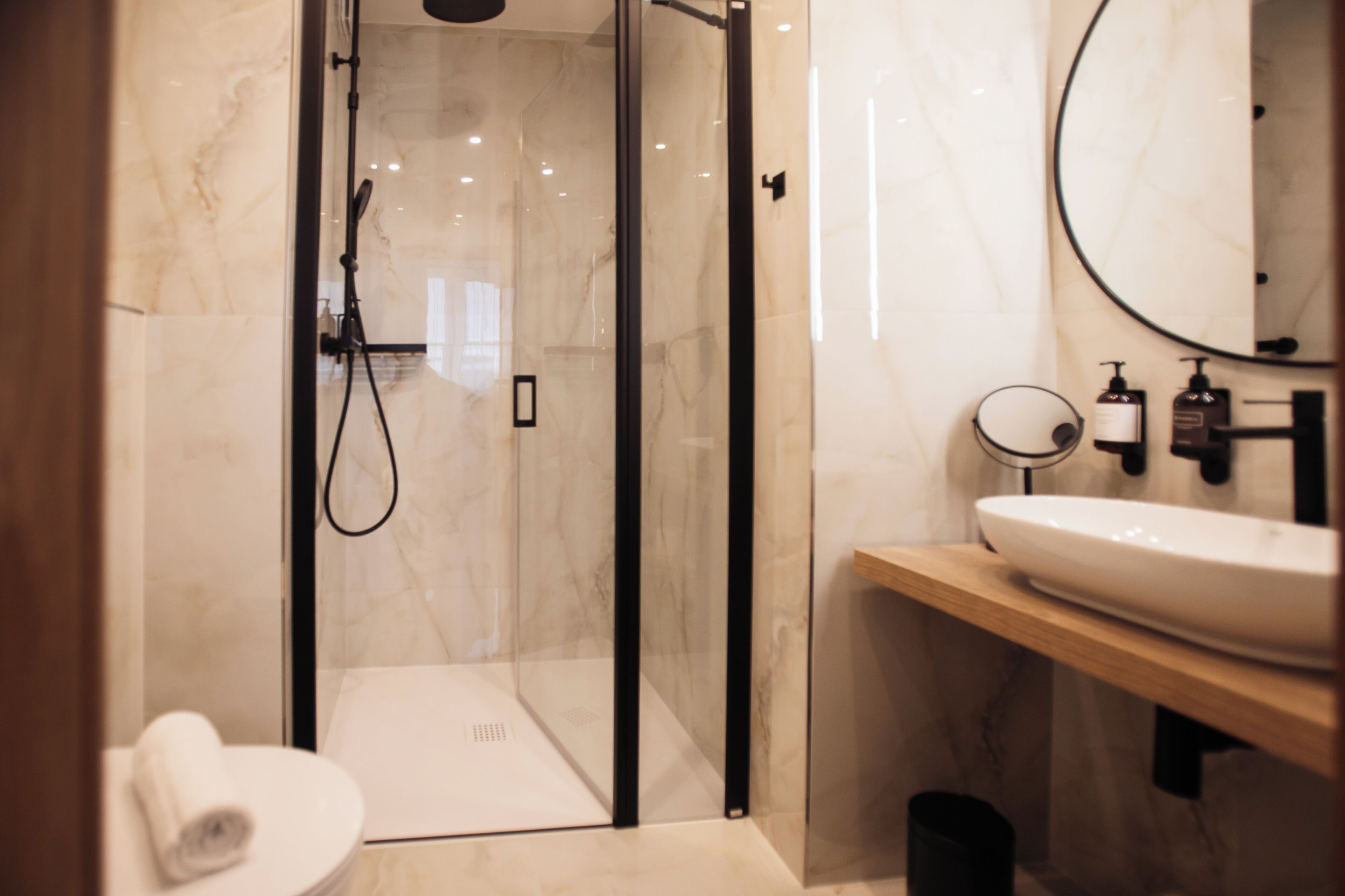 Un baño moderno con ducha envuelta en vidrio, inodoro blanco, tocador de madera con un espejo grande y lavabo blanco.