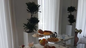 A buffet table with various food items and a three-tiered dessert display, surrounded by potted plants and white curtains.
