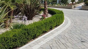 A two-story building with a tiled roof, a garden with various plants, and a paved driveway.