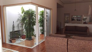 A living room with a sliding glass door leading to a balcony, wooden flooring, a brown sofa, and a kitchen area visible through the door. - Green House (Urasoe)