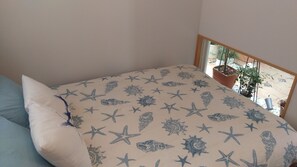 Ein Schlafzimmer mit einem Bett, das ein Meeresstern- und Muschelmuster zeigt, einem hellblauen Kopfkissen und Blick auf einen Balkon mit Blumentöpfen.