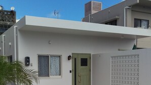 A modern two-story house with a green door, a small front yard with a patterned mat, and a white wall with a decorative panel. - Green House (Urasoe)