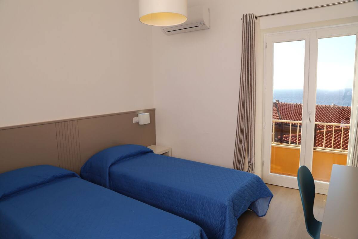 Habitación de hotel con dos camas, una ventana que ofrece vista a un tejado de tejas rojas y una pequeña mesa.