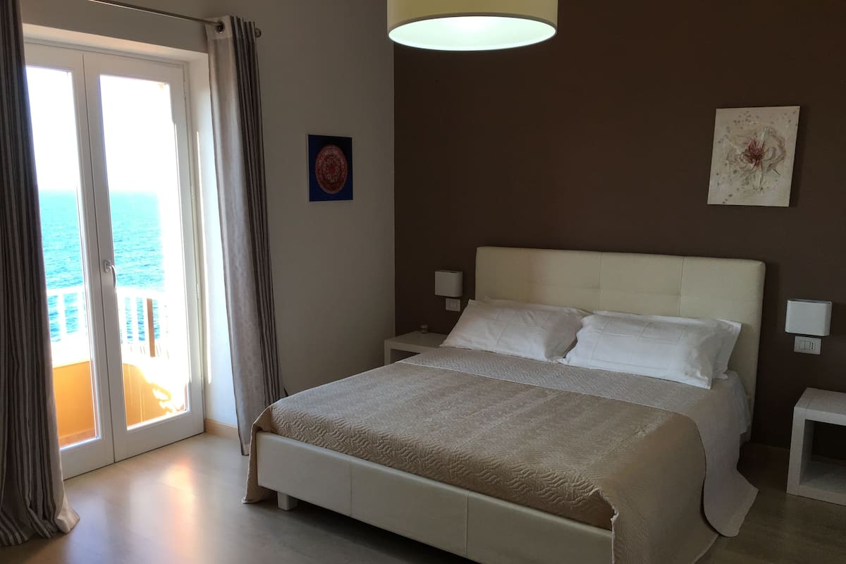 Un dormitorio con cama, mesita de noche, lámpara, un cuadro y vista al mar a través de una puerta corredera.