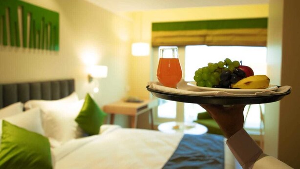 A hotel room with a bed, pillows, a bedside table, and a tray with fruit and a drink. - Regenta Central Harsha Vapi (Pardi)