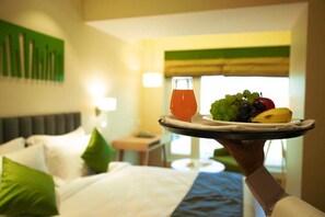 A hotel room with a bed, pillows, a bedside table, and a tray with fruit and a drink. - Regenta Central Harsha Vapi (Pardi)