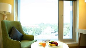 A green armchair with a dark cushion, a round white table with a fruit plate and a glass of orange juice, and a large window with a view of greenery and buildings. - Regenta Central Harsha Vapi (Pardi)