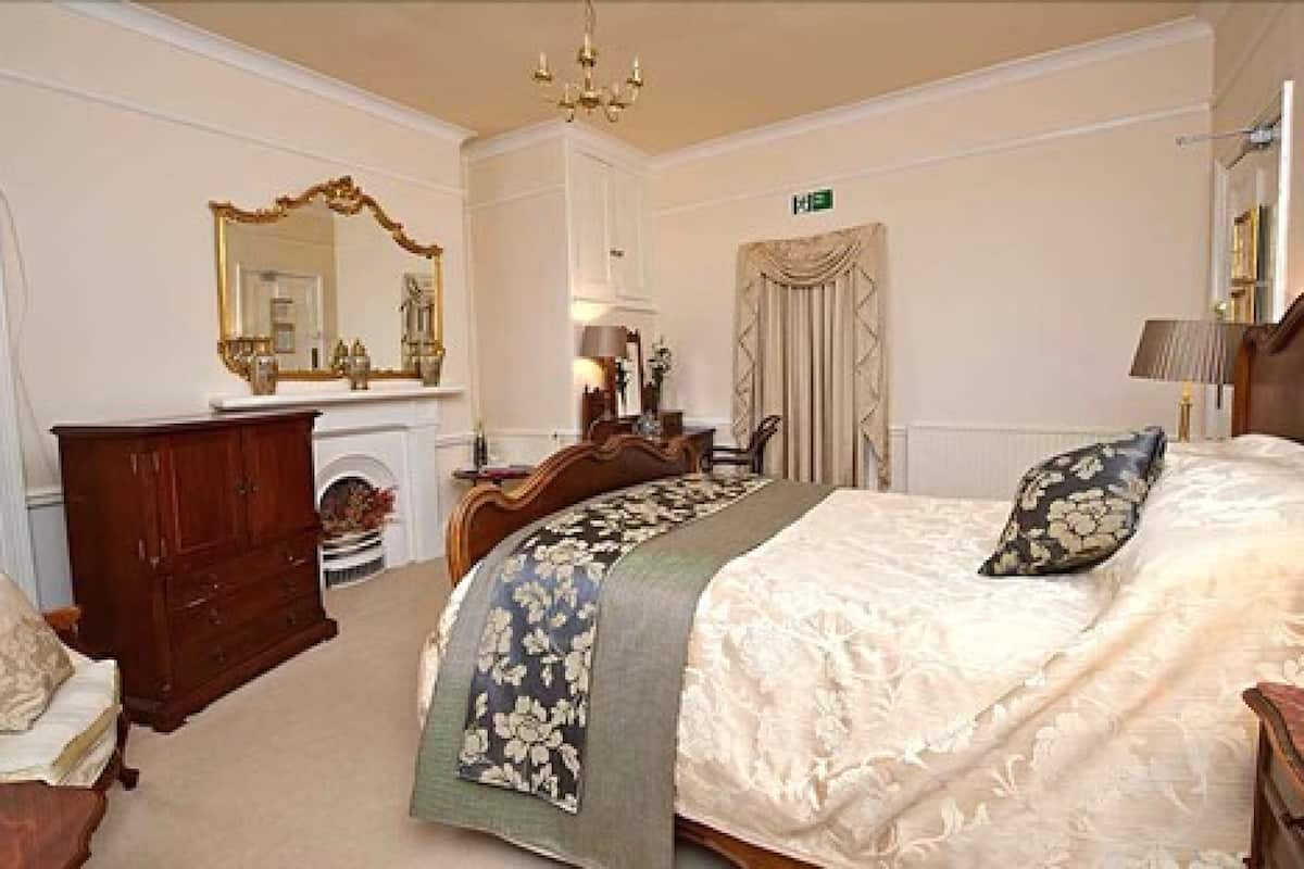 Une chambre d’hôtel avec un grand lit, une commode en bois, un miroir, une cheminée et un lustre.