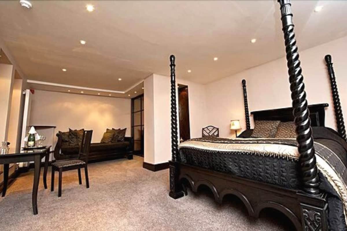 Un lit à baldaquin avec une tête de lit et un pied de lit sombres, un coin salon avec une table et des chaises, et un sol recouvert d’une moquette.