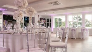 Un salón de banquetes con mesas redondas, sillas transparentes y centros de mesa florales blancos.