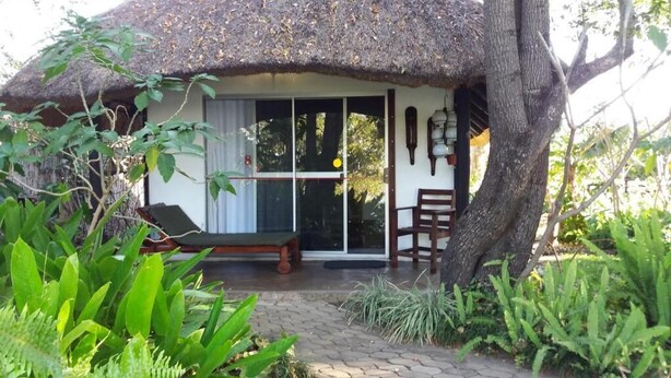 A thatched-roof hut with a bench and a chair outside, surrounded by lush greenery. - Caprivi River Lodge (Katima Mulilo)