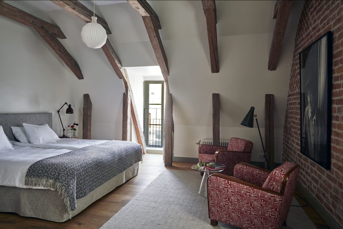 A bedroom with a bed, a red patterned armchair, a small table, and a fireplace.