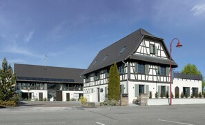 A modern building with a traditional half-timbered facade, a large glass entrance, and a parking lot in front. - Hotel Au Petit Pont (La Wantzenau)