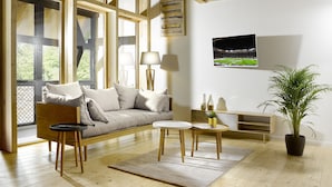 En hyggelig stue med træloft, store vinduer, en sofa, et sofabord, en lampe, et fjernsyn og en potteplante.