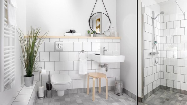 Un baño moderno con inodoro, lavamanos y ducha. Hay una planta en maceta, un perchero para toallas y un taburete pequeño.
