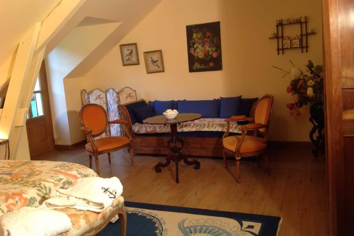 Une pièce chaleureuse avec un plancher en bois, un canapé avec des coussins bleus, deux chaises en bois, une petite table avec un bol, et un lit recouvert d’une couvre-lit à motifs floraux.