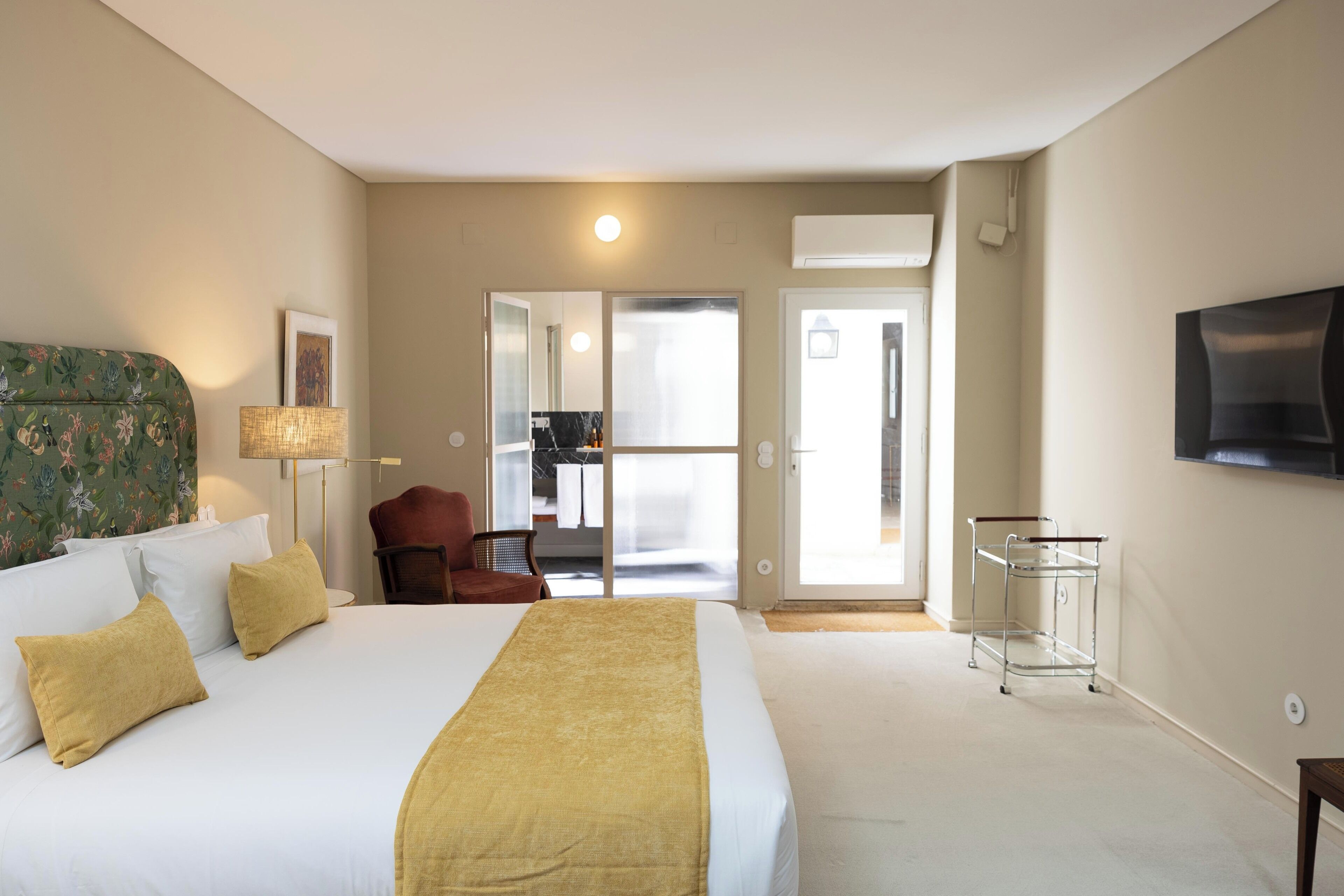 A hotel room with a large bed, a flat-screen TV mounted on the wall, a chair, a lamp, and a sliding door leading to a balcony.