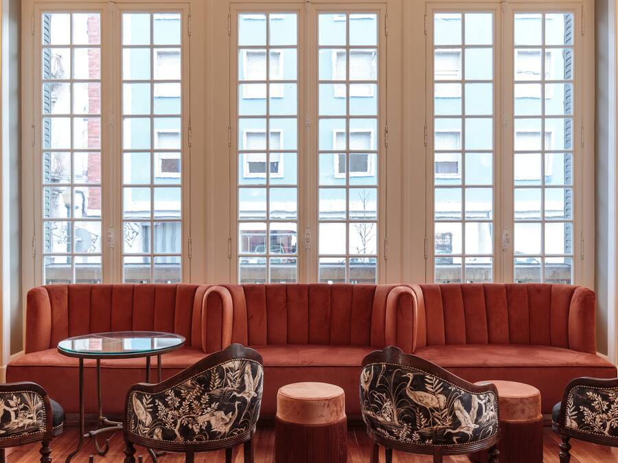 A room with a red sofa, two ornate chairs, a small round table, and large windows with a view of buildings.