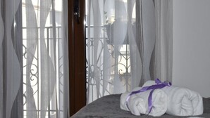 Una cama con una toalla blanca enrollada atada con un lazo morado, una puerta de madera y un balcón con barandilla de metal y cortinas traslúcidas.
