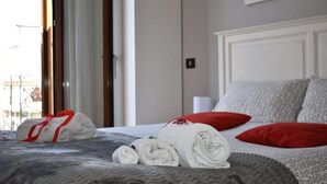 Una cama bien hecha con toallas enrolladas y una almohada roja, una ventana con cortinas, y una pared con un cuadro enmarcado.