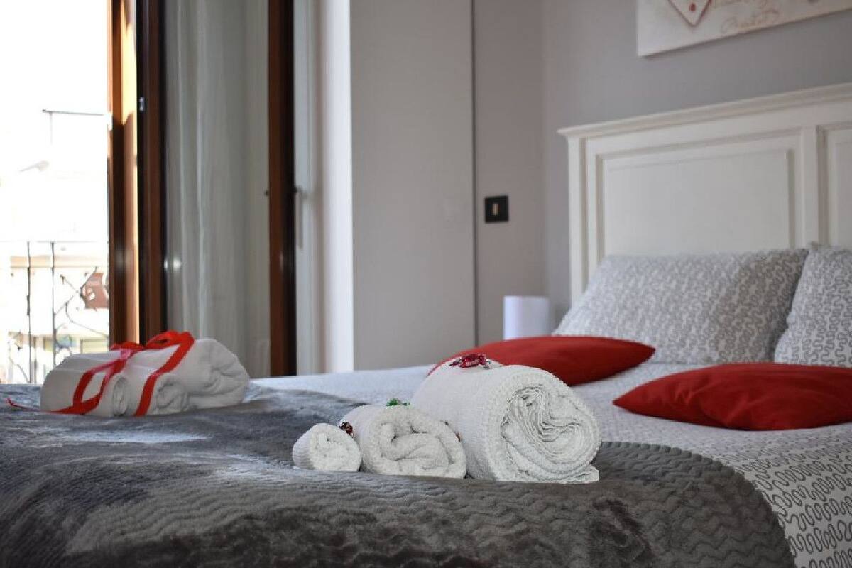 Un lit bien fait, avec des serviettes pliées et un coussin rouge, une fenêtre avec des rideaux et un mur orné d’un tableau encadré.