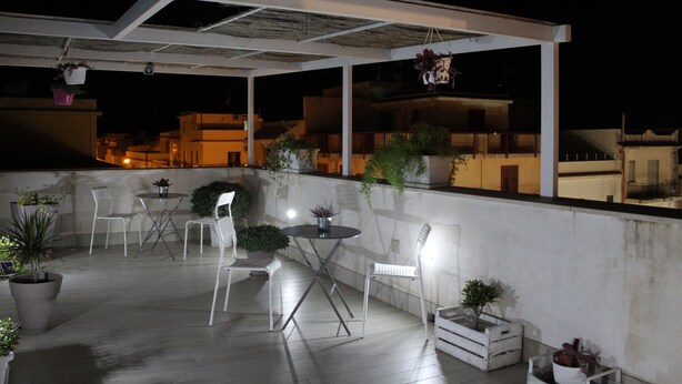 Una terraza en la azotea con mobiliario exterior, plantas en macetas y vistas de la ciudad de noche.