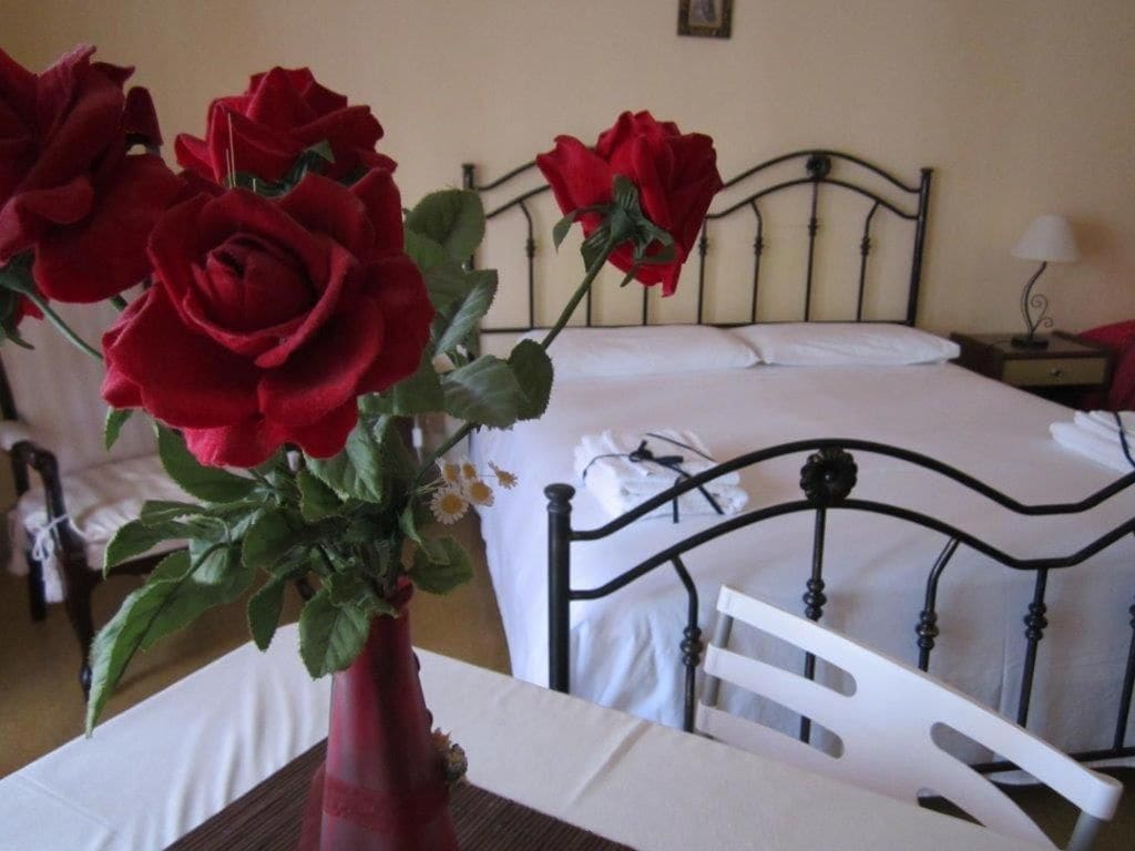 A room with a bed, a white chair, and a table with a vase of red roses.