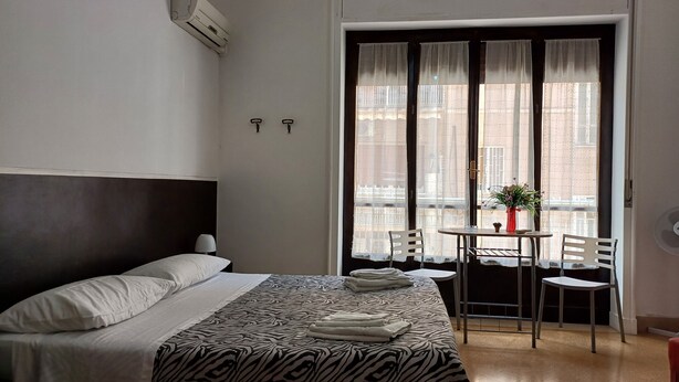 A hotel room with a bed, a small table with chairs, a window with curtains, and an air conditioning unit. - Da Mary Guest House (Palermo)