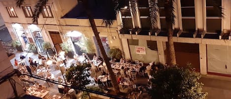 An outdoor dining area with multiple tables and chairs, surrounded by buildings and palm trees.