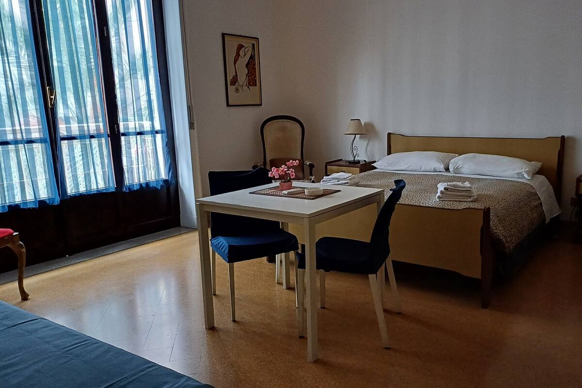 Habitación de hotel con cama, mesa con sillas, escritorio, silla, mesita de noche, lámpara, un cuadro en la pared y ventana con cortinas.