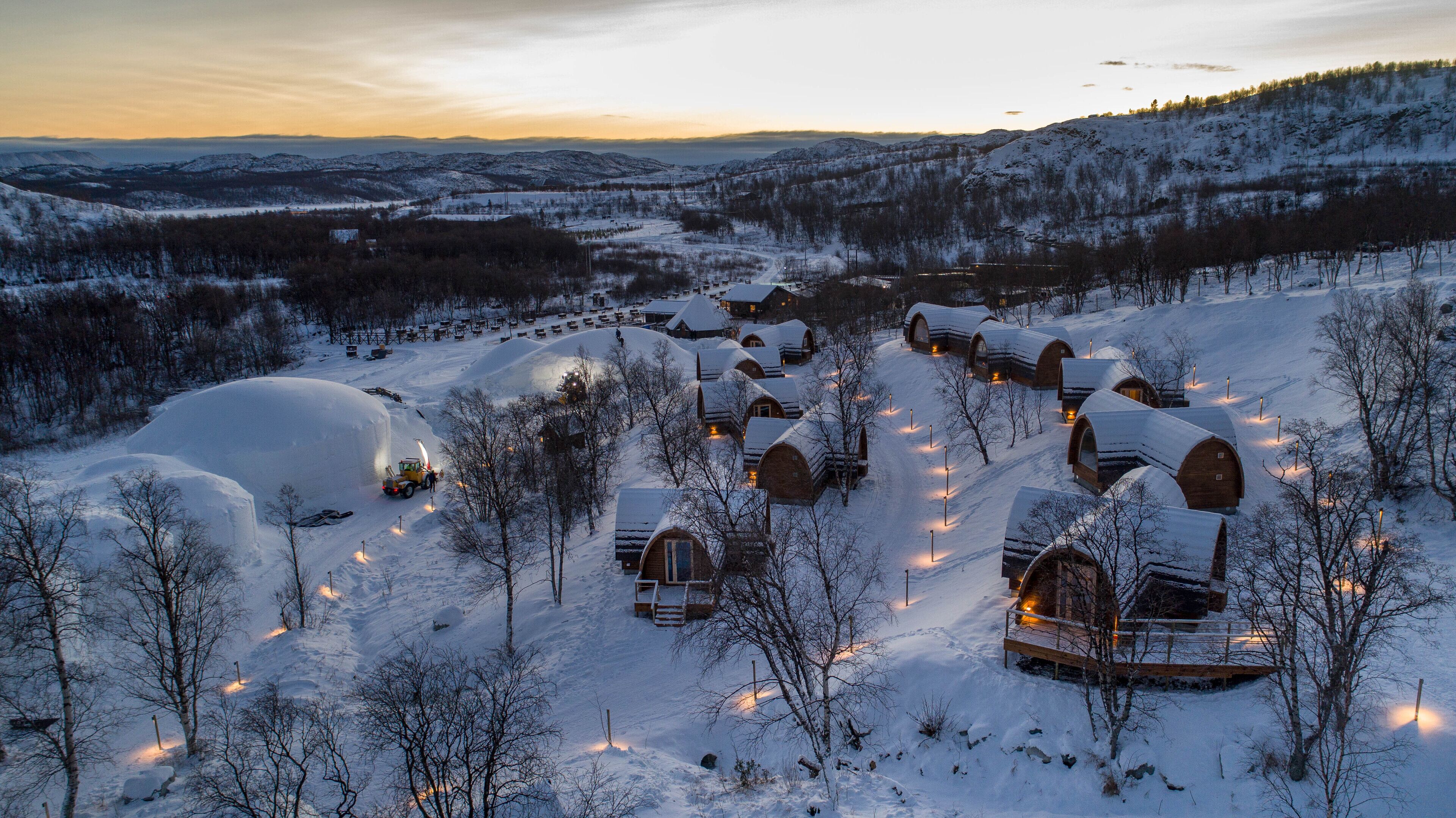 A snow-covered landscape with several wooden cabins arranged in a semi-circle, surrounded by trees and hills.