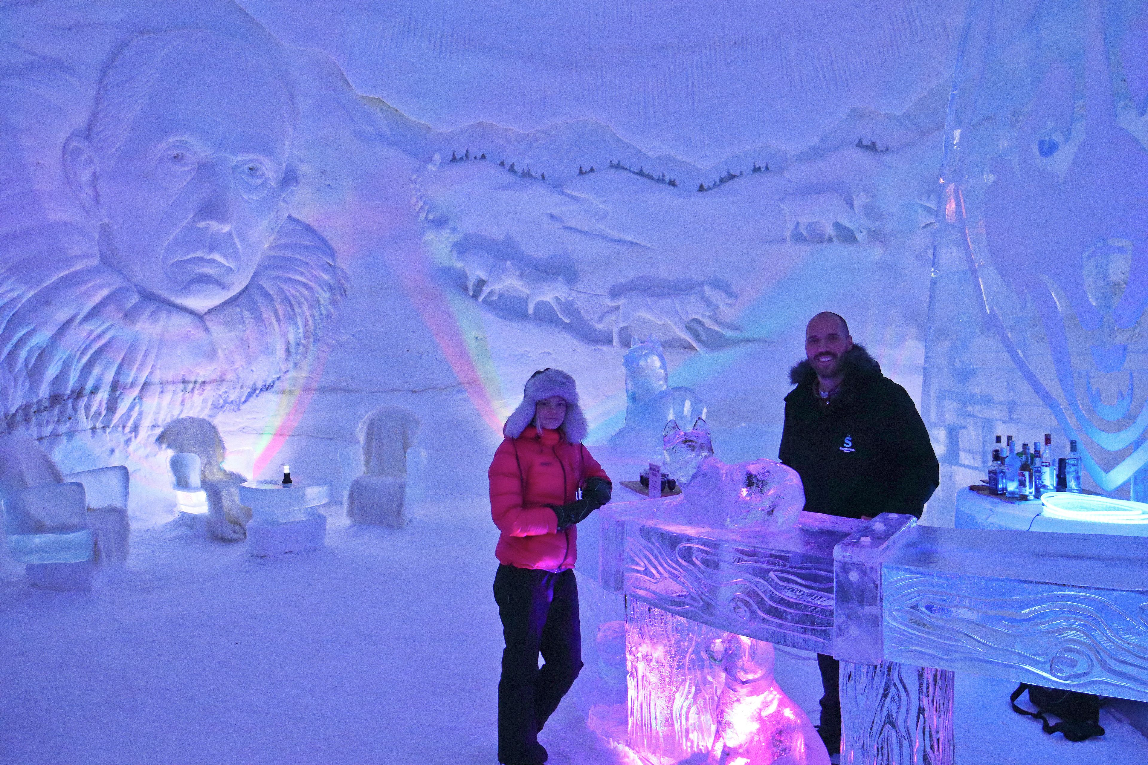 An ice bar with a bartender, ice sculptures, and a mural in a snowy setting.