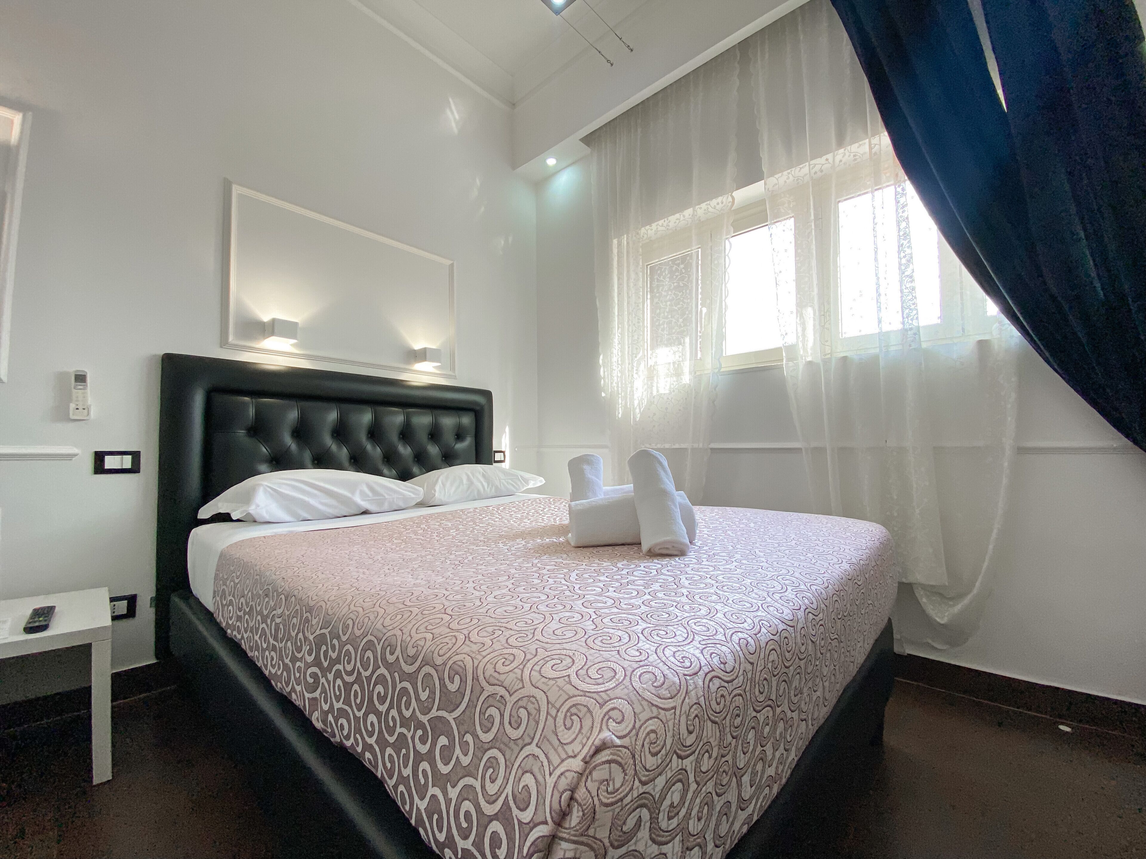 Un lit bien fait, recouvert d’une couvre-lit à motifs, une table de chevet avec une télécommande et une fenêtre ornée de rideaux légers.