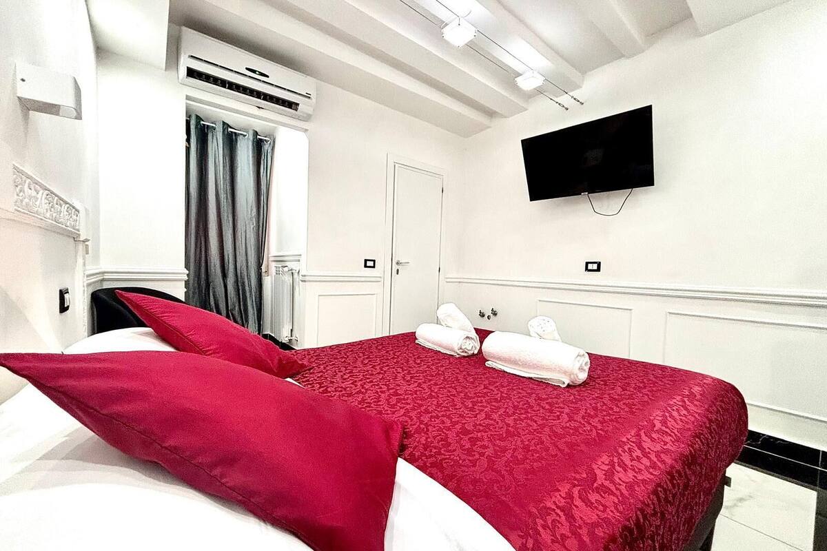 A hotel room with a bed, a flat-screen TV mounted on the wall, a window with curtains, and an air conditioning unit.