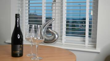 A bottle of wine and two glasses on a wooden table by a window with blinds.