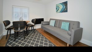 A modern living room with a grey sofa, black and orange chairs, a patterned rug, a wall-mounted artwork, and a ceiling light. - The Plough Retreat (Newark)