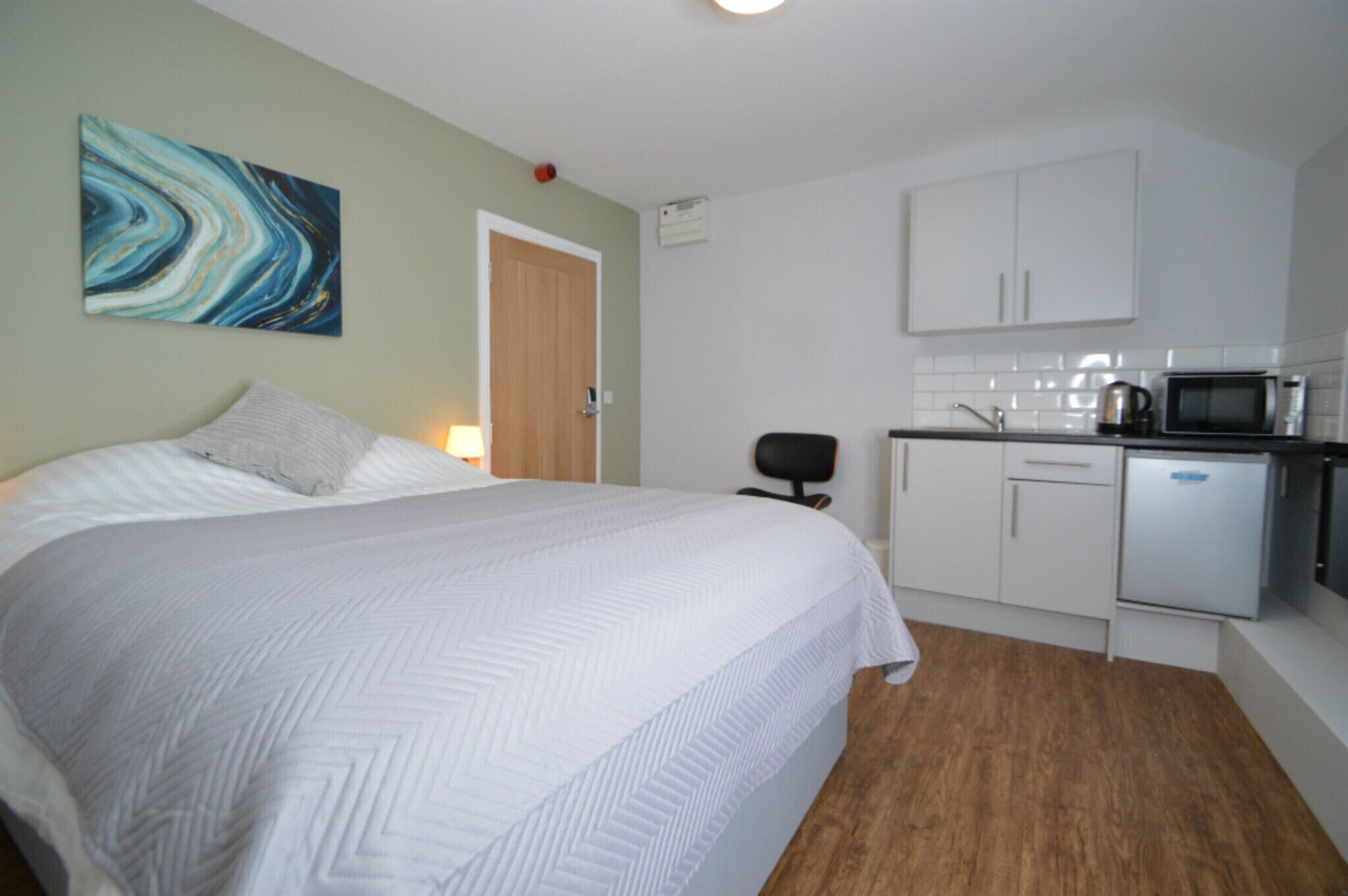 Een slaapkamer met een groot bed, een nachtkastje en een nachttafel. Er is een kleine kitchenette met een magnetron, waterkoker en een koelkast.