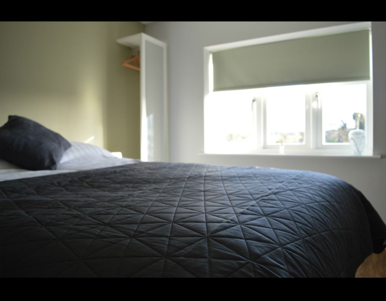 A bed with a dark quilted bedspread, a window with blinds, and a view of the outside.